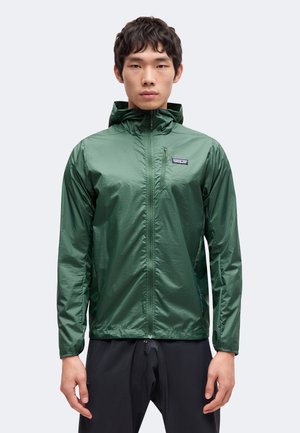 Mann trägt eine grüne Kapuzenjacke mit Reißverschluss und schwarze Hose, steht vor einem schlichten hellen Hintergrund.