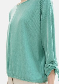 Žena v tyrkysové a bílé tenké pruhované blůze s nabíranými a uvázanými manžetami, zkombinované se světle béžovými kalhotami.