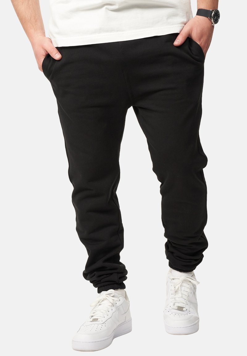 Black sweatpants with a relaxed fit, tapered ankles, and side pockets, paired with white sneakers for a casual look.