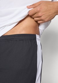 Fekete sportnadrág fehér oldalsó panellel, rugalmas derékrésszel és puha, texturált anyagból. Egy kéz felemel egy fehér pólót a deréktájékon.