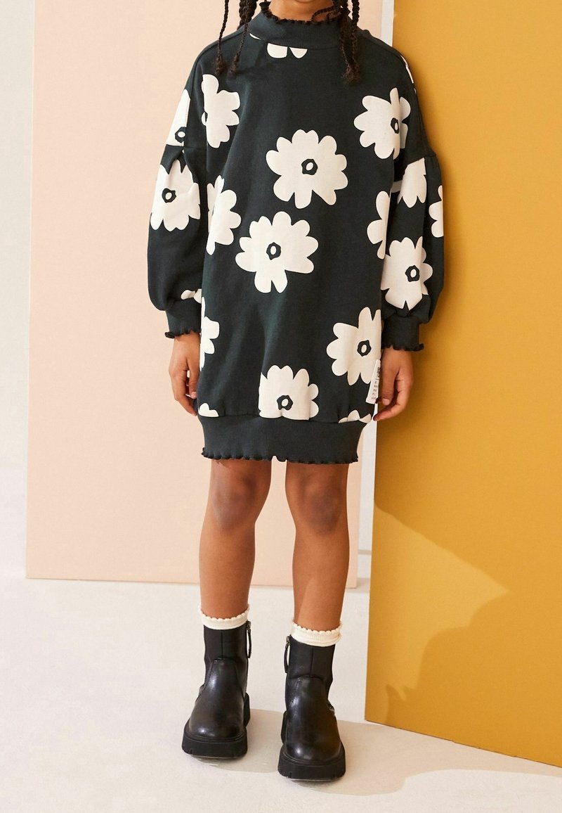 Czarna sukienka swetrowa z białym wzorem kwiatowym i bufiastymi rękawami. Zawiera falbaniasty dół. Noszona z czarnymi botkami za kostkę z falbaniastym mankietem.