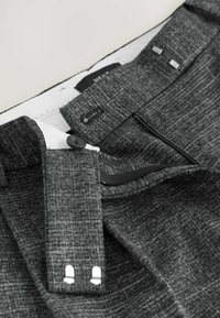Pantaloni grigi scuri su misura con la cerniera aperta e il bottone di chiusura slacciato, che mostrano la fodera bianca interna e i ganci metallici in vita.