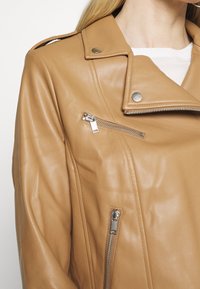 Veste de biker en cuir tan avec des détails en métal argenté, des boutons-pression sur le col et une texture lisse. Une poche zippée sur la poitrine.