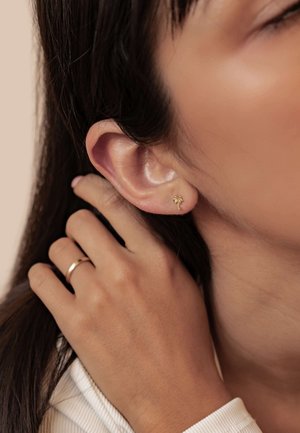 Goldene Steckerohrringe in Form von Palmenblättern am Ohr eines Modells mit glatter Textur; Hautton und ein goldener Ring an der Hand, die das Haar berührt.