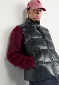 Ung man med vitiligo som bär en rosa keps, burgunderfärgad fluffig tröja och en glänsande svart pufferjacka med händerna i fickorna mot vit bakgrund.