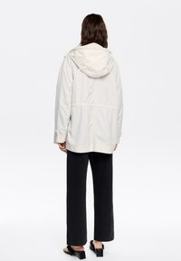 Hellbeige Jacke mit Kapuze, elastischem Bund und weicher Textur. Kombiniert mit schwarzen weit geschnittenen Hosen und Absatzsandalen mit Fransenakzenten.