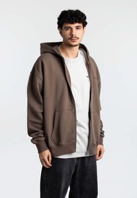 Hoodie marrone con zip, dotato di un ampio cappuccio, tasche frontali e polsini a costine, indossato sopra una maglietta grigia chiara e abbinato a jeans scuri.