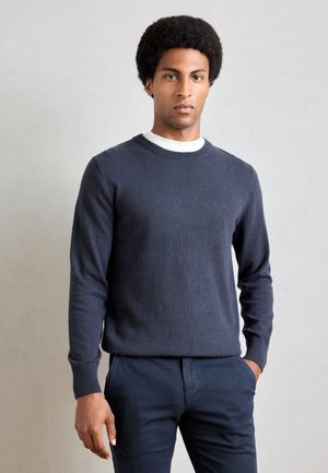 Navyblauwe gebreide trui met een ronde hals en lange mouwen, gedragen over een wit overhemd. Gladde textuur, slim fit, geen opvallende logo's.