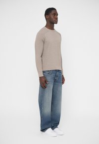 Długi sweter z długim rękawem w jasnobeżowym kolorze o prążkowanej fakturze, zestawiony z luźnymi niebieskimi dżinsami i białymi trampkami. Model stoi na jednolitym tle.