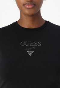 Črna bombažna majica s krojnim ovratnikom, kovinskim zlatim logotipom "GUESS" in trikotnim poudarkom pod "originals." Gladka tekstura.