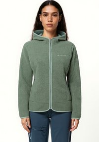 Giovane donna che indossa una giacca in pile verde con zip e cappuccio e pantaloni blu, in piedi su uno sfondo bianco.