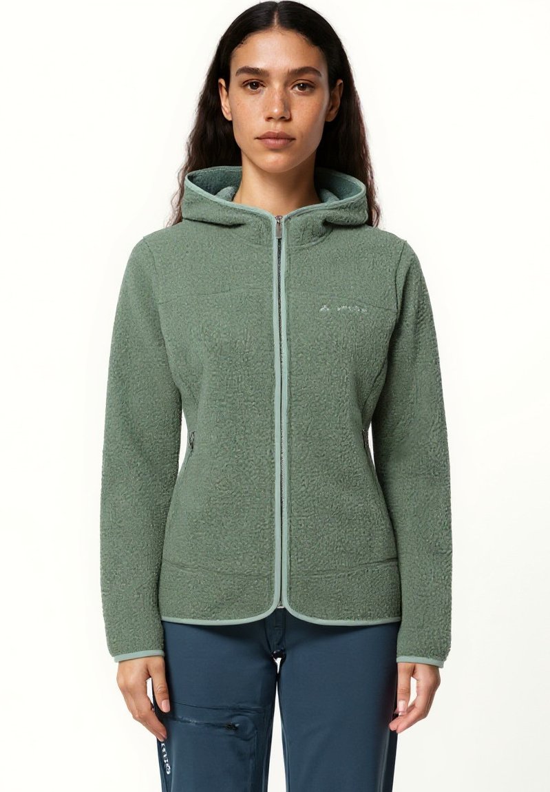 Giovane donna che indossa una giacca in pile verde con zip e cappuccio e pantaloni blu, in piedi su uno sfondo bianco.