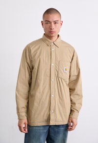 Striped, button-up shirt in beige with thin blue lines. Features a collar, single chest pocket, and white buttons. Soft cotton texture.