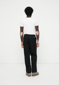 Person with vitiligo on arms wearing white T-shirt, black cargo pants, and beige boots, standing facing a white wall.
