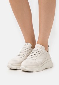 Les baskets en cuir blanc présentent une semelle épaisse, un bout arrondi et un design minimaliste. Les détails comprennent des lacets texturés et un logo sur la languette.