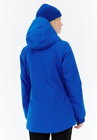 Chaqueta impermeable azul con capucha, que presenta puños elásticos y una textura suave. Lleva un gorro a juego, todo sobre un fondo blanco.