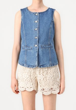 Vrouw die een mouwloze blauwe denim blouse met knopen draagt en crèmekleurige korte broek met bloemenkant, staand tegen een effen achtergrond.