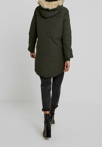 Grön parkas med en foderhuva i päls, långa ärmar och en böjd fåll, tillsammans med mörka jeans och ankelstövlar. Slät tygtextur.