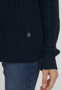Maglione lavorato a maglia di un blu navy scuro con un motivo testurizzato, polsini e orlo a coste. Presenta un piccolo logo metallico vicino alla cucitura inferiore.