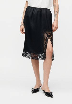 Donna che indossa una gonna di raso nera con bordatura in pizzo e spacco laterale, top bianco e scarpe nere con tacco basso a punta con dettaglio metallico.