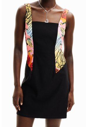 Kobieta ubrana w czarną sukienkę bez rękawów z kolorowymi abstrakcyjnymi wzorzystymi bocznymi panelami oraz srebrnym naszyjnikiem w kształcie serca.