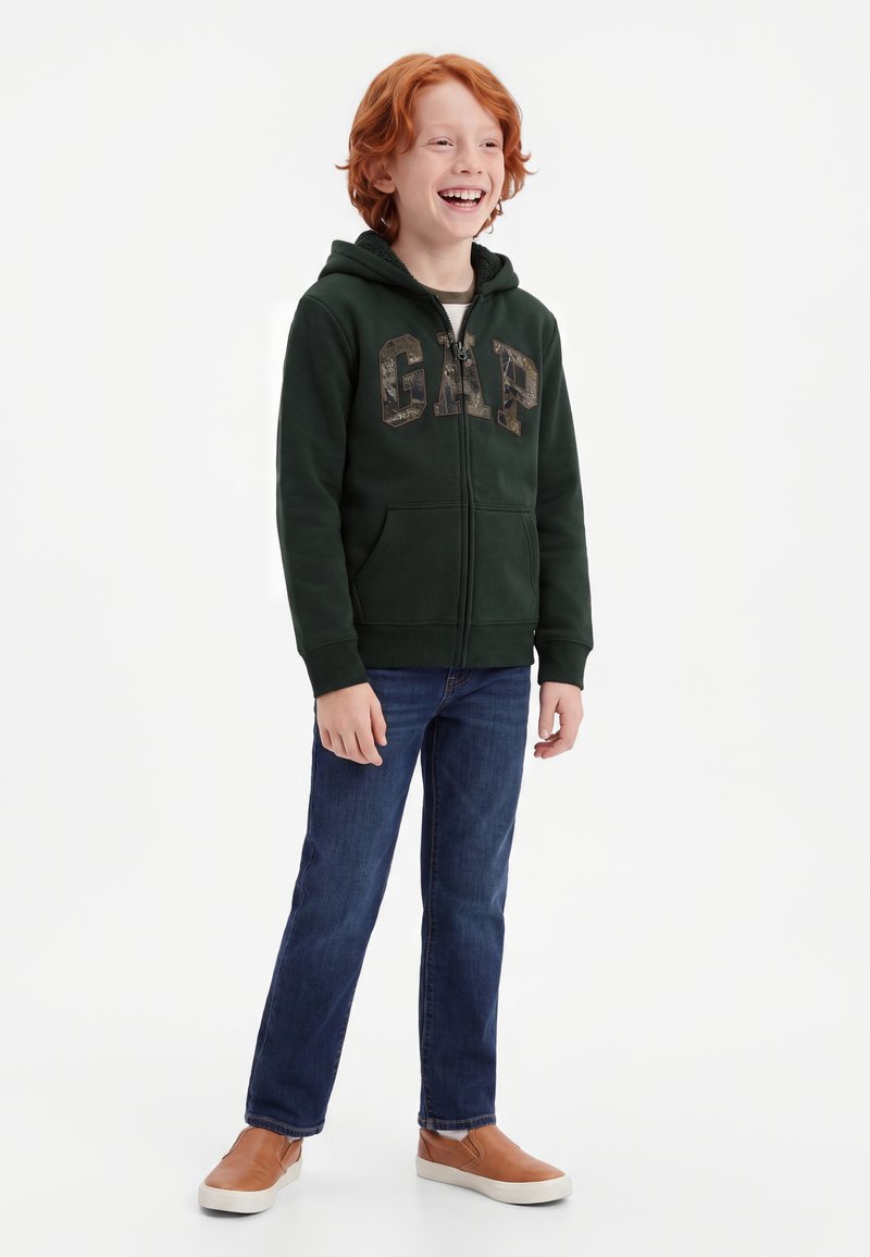 Garçon aux cheveux roux souriant, portant un sweat à capuche zippé vert foncé avec le logo GAP, un jean bleu et des chaussures marron sans lacets, debout sur fond blanc.