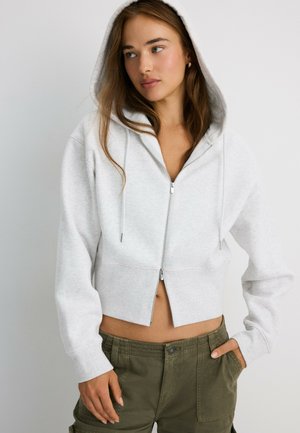 Jeune femme portant un sweat à capuche court gris clair zippé avec la capuche mise, associé à un pantalon cargo vert, regardant sur le côté.