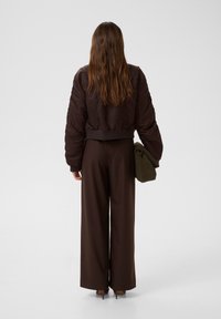 Jachetă bomber căptușită, maro, asociată cu pantaloni largi, de culoare maro. Modelul ține în mână o geantă pliabilă, verde. Părul lung îi cade pe spate.