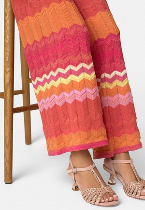 Benedenbenen met een multicolor zigzag gebreide broek en roze T-strap sandaaltjes met studs en hakken, zittend op een houten stoel met zichtbare poten.