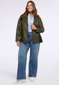 Leende kvinna i olivgrön läderjacka, ljusblå skjorta med knappar, vida blå jeans och vita sneakers som står på vit bakgrund.