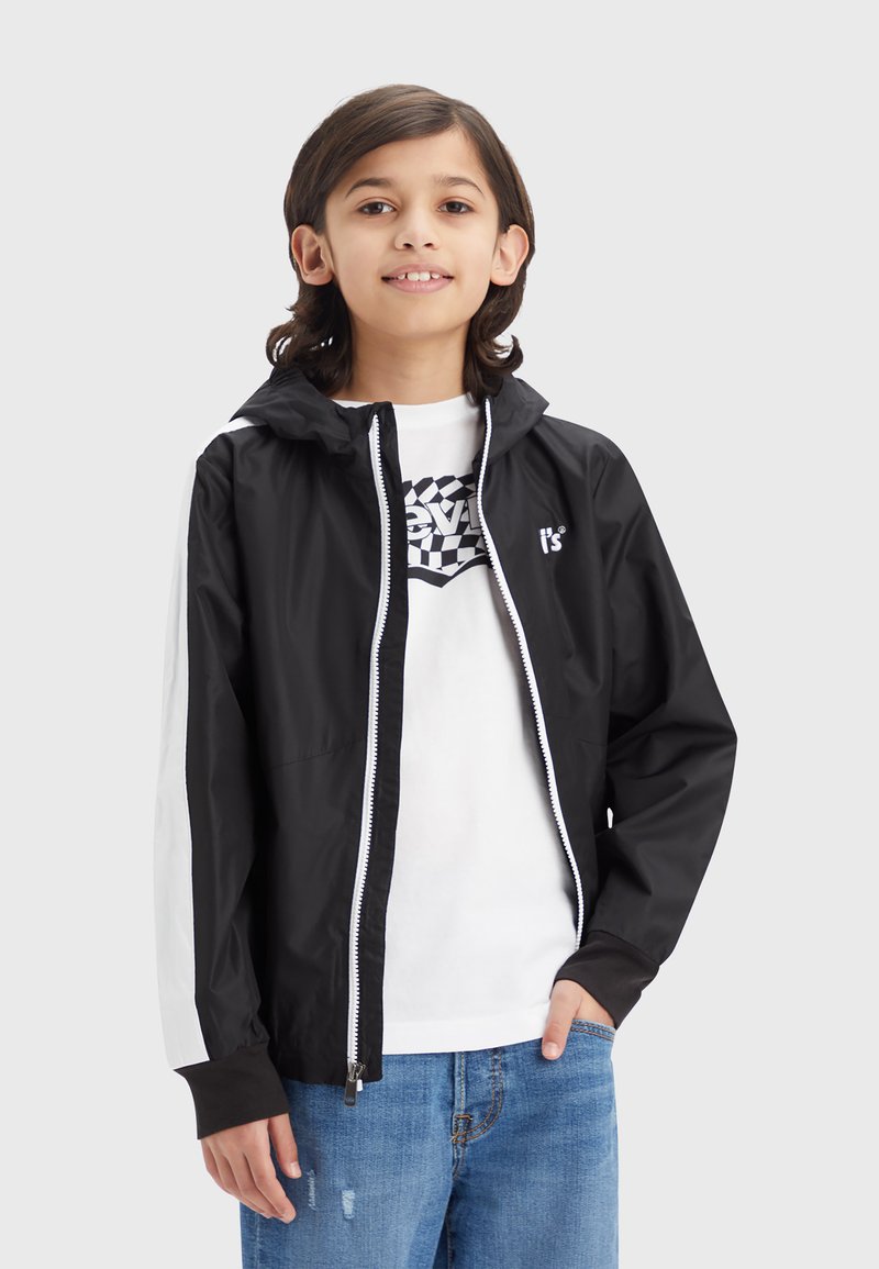 Schwarze leichte Zip-Jacke mit Kapuze, ausgestattet mit weißen Seitenstreifen und einem weißen Reißverschluss. Getragen über einem weißen T-Shirt mit grafischem Design.