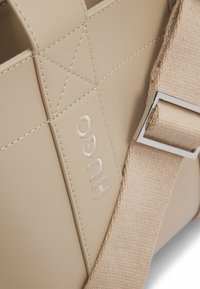 Beige leather handbag featuring stitched detail and a silver logo. The strap is made of fabric with a metallic buckle.