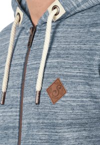 Sudadera azul jaspeada con cremallera, cordón crema y parche de logo de cuero. Presenta ojales de metal y tejido texturizado.