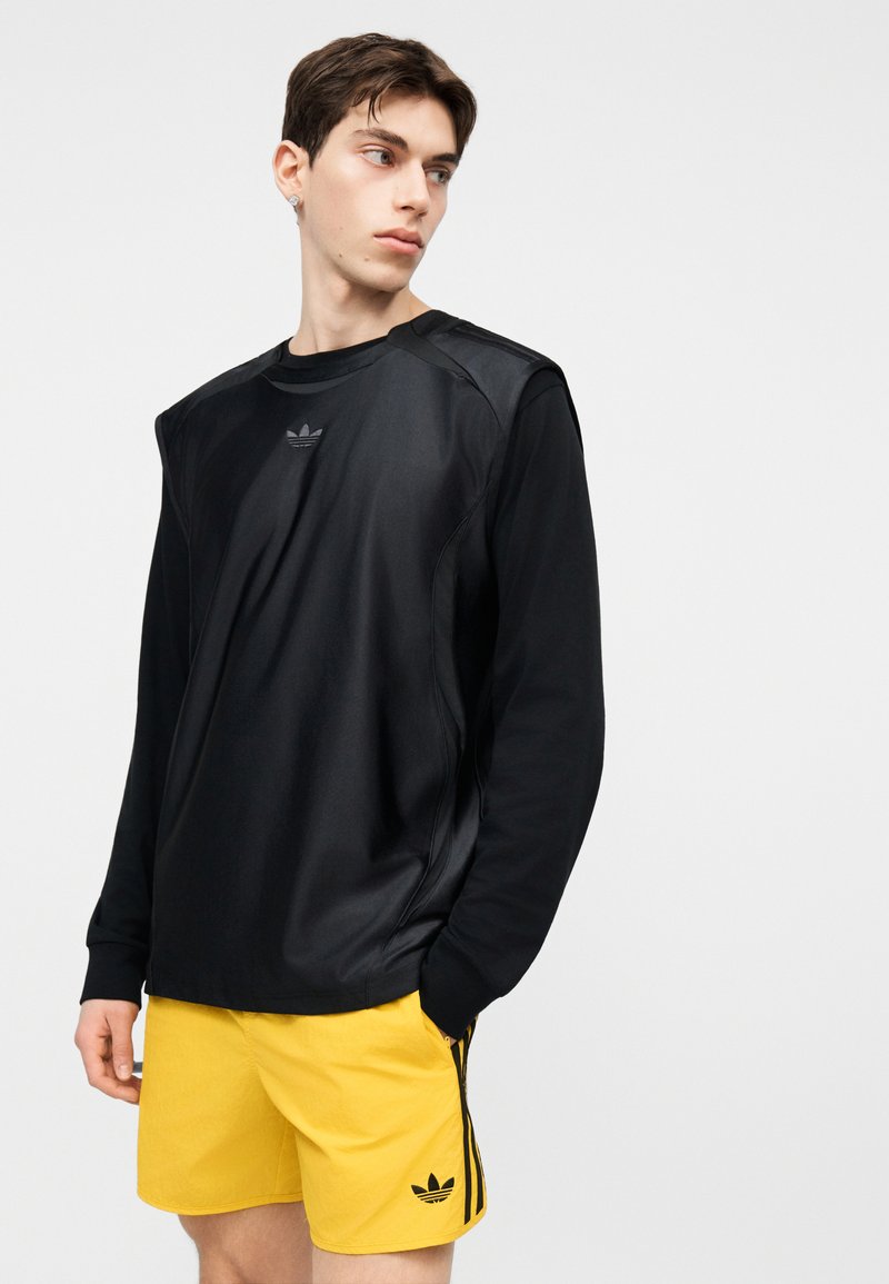 Unge mand iført sort langærmet skjorte og gule atletikshorts med sort logo, kiggede til venstre med hånden i lommen.