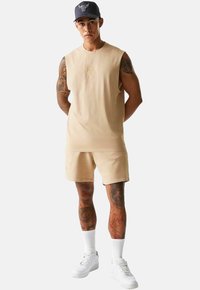 Top in cotone beige senza maniche e pantaloni corti abbinati. Presenta un logo ricamato. Completato da sneaker bianche e un cappellino blu.