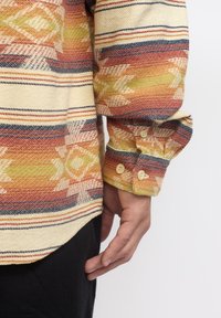 Hand van een persoon naast de manchette van een gestreepte lange mouw shirt met beige knopen in oranje, geel, marineblauw en crème kleuren en geometrische ontwerpen.