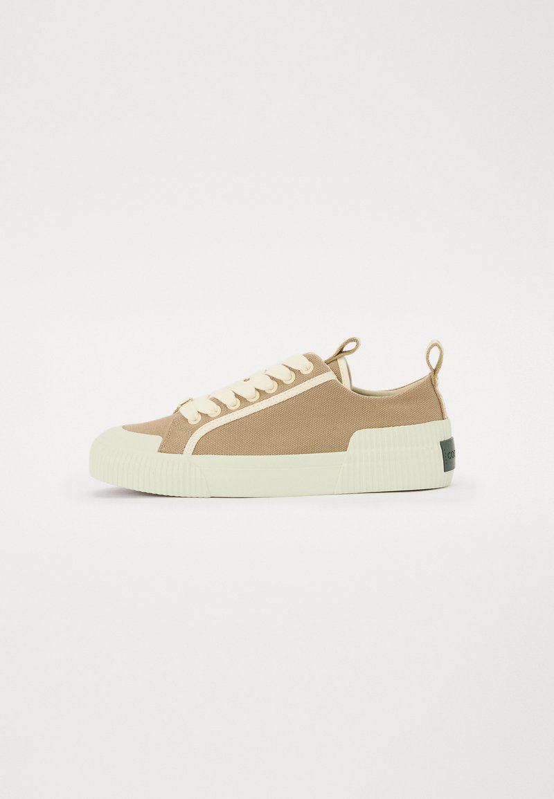 Baskets en toile beige avec une semelle en caoutchouc crème texturée, des lacets blancs et une languette arrière dotée d'un petit logo.