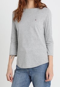 Grå långärmad skjorta i mjukt tyg med båthals. Har en liten logotyp på vänster bröst och en avslappnad passform. Kombinerad med blå jeans.