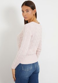 Suéter de punto cableado en rosa claro con cuello redondo y dobladillo acanalado, con mangas largas y detalles de patrón texturizado. Combinado con jeans azules.