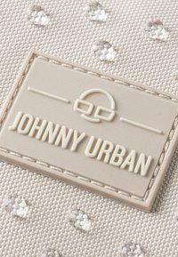 Kremowa powierzchnia materiału z teksturowym splotem, z wystającą beżową etykietą z napisem "JOHNNY URBAN" oraz logo. Widoczne są małe, przezroczyste ozdoby.