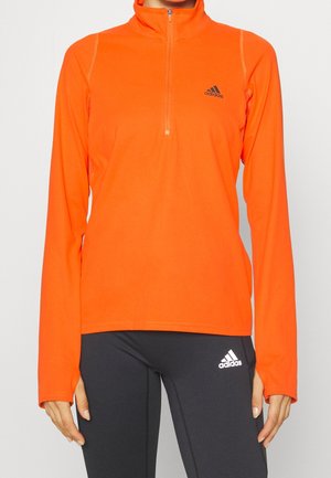 Haut de sport orange à manches longues avec fermeture éclair, associé à un legging noir, tous deux avec logos Adidas visibles sur la poitrine et la cuisse.
