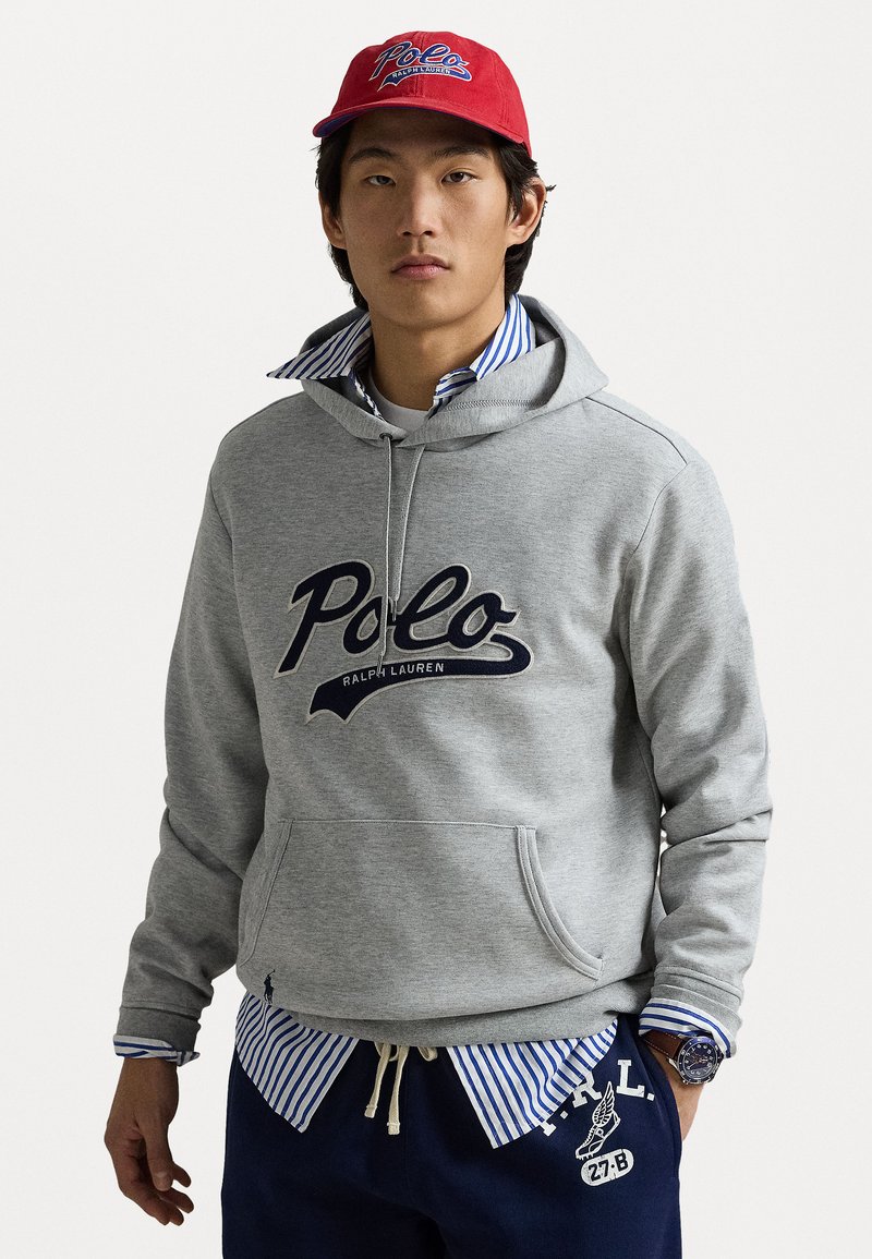 Felpa grigia con grande logo "Polo Ralph Lauren", indossata sopra una camicia a righe blu e bianche con colletto. Abbinata a pantaloni della tuta navy e un cappellino rosso.