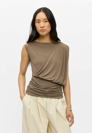 Femme aux longs cheveux noirs portant un haut taupe drapé sans manches et un pantalon ample de couleur crème, debout les mains dans les poches.