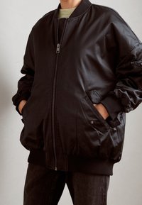 Blouson aviateur noir à finition lisse, fermeture éclair, poignets et ourlet côtelés, poches latérales, et coupe décontractée. Tissu texturé apportant de la profondeur.