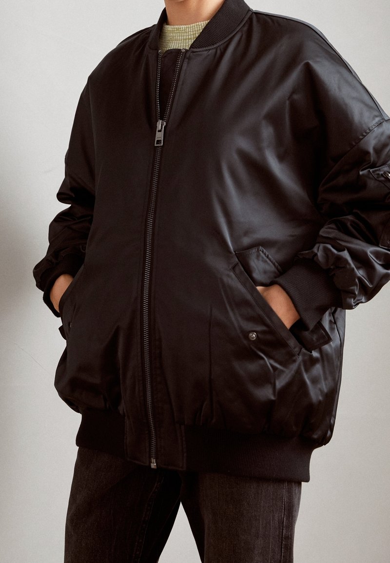 Blouson aviateur noir à finition lisse, fermeture éclair, poignets et ourlet côtelés, poches latérales, et coupe décontractée. Tissu texturé apportant de la profondeur.