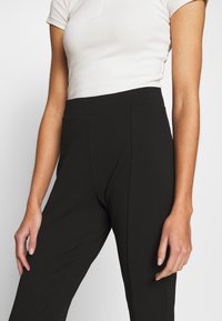 Černé kalhoty s vysokým pasem z hladké látky s přizpůsobeným střihem; mají přední záhyby a široké nohavice. Nošeny s obtaženým bílým topem.