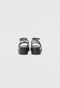 Sandales noires avec un talon épais, dotées de fines lanières et de boucles réglables à la cheville. Texture lisse avec un design minimaliste.