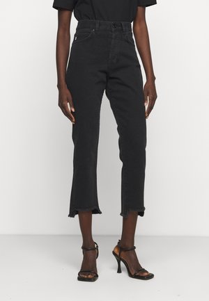 Personne portant un jean noir court taille haute avec des ourlets effilochés et des sandales noires à talons hauts avec des brides, debout devant un fond uni.