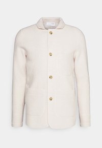 Jasnobeżowa kurtka z wełny z kołnierzem, zapięciem na guziki z przodu, dwoma naszywanymi kieszeniami oraz fakturowanym wykończeniem tkaniny.