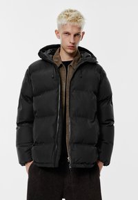 Črna puhasta jakna s kapuco, s prešitimi predeli, sprednjo zadrgo in potegalko. Nošena preko rjave jopice z zadrgo.